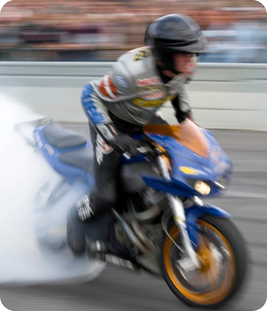 Un motard casqué effectue un burnout à grande vitesse sur une moto bleue, créant un gros nuage de fumée. Une foule floue observe la scène en arrière-plan.